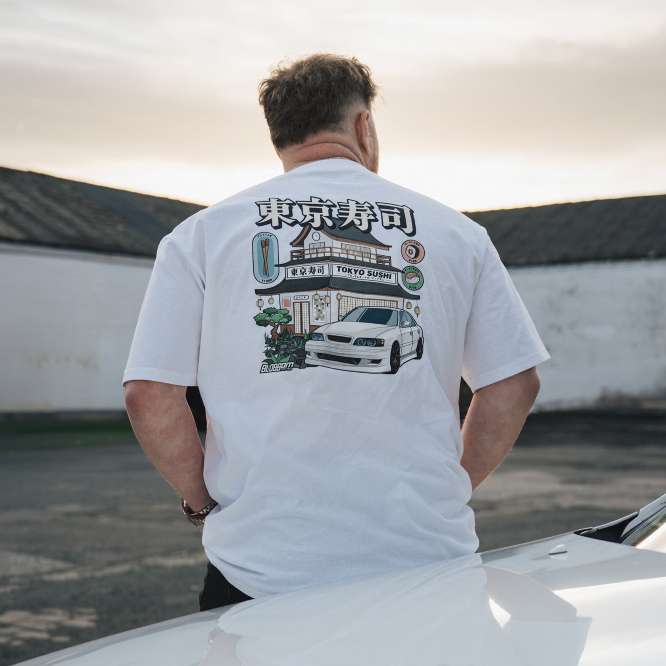 A person wearing a white crewneck t-shirt with a graphic design on the back, featuring a sushi restaurant and a car, standing with their back towards the camera.