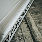 A close-up of the Blossom Automotive "Blossom URL - XL Die-Cut Sticker (x1)" on a white car side skirt, showing part of the website address beside a concrete surface.