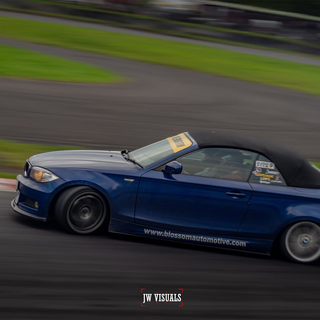 A blue convertible races around a track, showcasing Blossom URL - XL Die-Cut Sticker (x1) by Blossom Automotive on the window and hood. JW Visuals logo is visible at the bottom, with motion blur in the background.