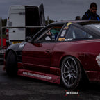 A maroon drift car with racing decals and low suspension is parked near a white van at an outdoor event. The Blossom Automotive logo appears in the corner, highlighting their Blossom URL - XL Die-Cut Sticker (x1). Two people stand in the background.