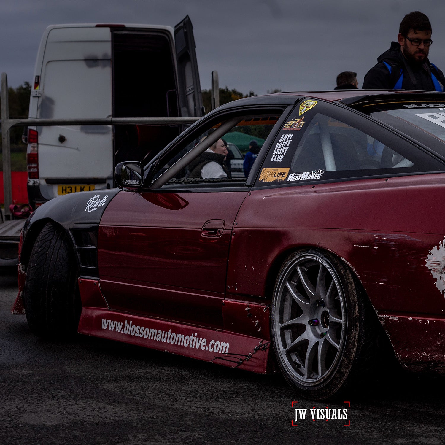 A maroon drift car with racing decals and low suspension is parked near a white van at an outdoor event. The Blossom Automotive logo appears in the corner, highlighting their Blossom URL - XL Die-Cut Sticker (x1). Two people stand in the background.