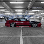 A lowered red and black sports car with aftermarket wheels, decals, and a large rear wing is parked in an empty parking garage. Its side skirt displays the Blossom Automotive "Blossom URL - XL Die-Cut Sticker (x1).