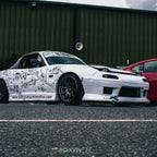 A white sports car featuring Blossom Automotive’s Blossom URL - XL Die-Cut Sticker is parked on asphalt next to a red vehicle, in front of a green industrial building. Photo by @Ducky_17.