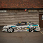 A Blossom Automotive sports car featuring a Blossom URL - XL Die-Cut Sticker is parked in front of a wooden building. The white car has blue and silver graphics, sponsor logos, custom wheels, and a body kit.