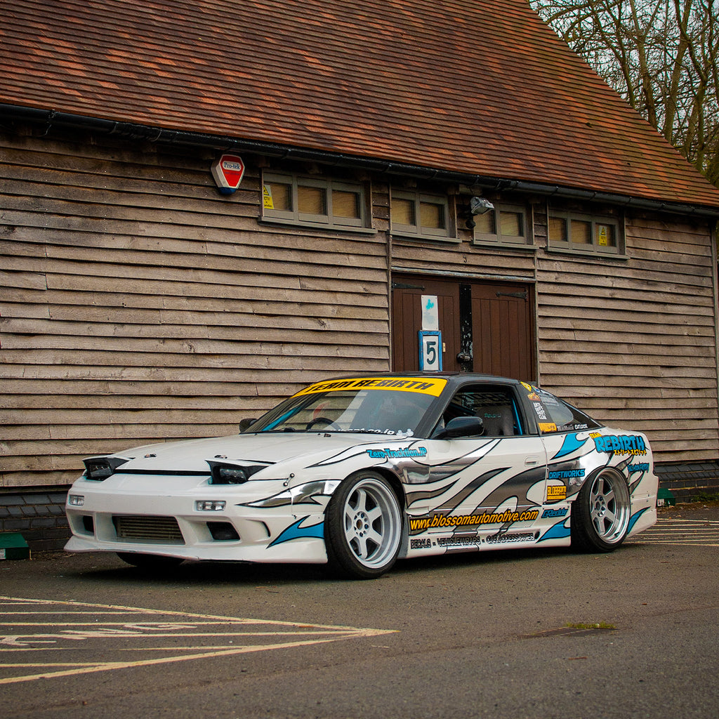A Blossom Automotive "Blossom URL - XL Die-Cut Sticker (x1)" is featured on a modified white sports car with blue and gray graphics, wide body kit, lowered suspension, and racing decals parked by a wooden building with a slanted roof.