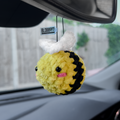 The Squishy Bumblebee - Car Mirror Hanger by Blossom Automotive, a cute yellow and black plush with white wings and pink cheeks, dangles from a car’s rearview mirror. A blurred outdoor view with greenery appears in the background.