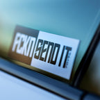 A Blossom Automotive FCKN Send It - Slap Sticker in black and white is angled on a car window, showing off its bold text with a blurred background seen through the glass.