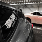 Close-up of a car window showing the Blossom Automotive “Good Vibes Only - Slap Sticker” with waves, clouds, and “Kelsea S. Astra.” A white car is parked beside it in a covered parking area.
