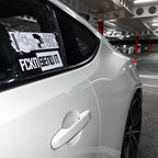 A white sports car is parked in an indoor garage. On the rear side window are two stickers: Blossom Automotive’s “Good Vibes Only - Slap Sticker” with a wave illustration, and another that says FCKNSENDIT.