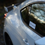 Close-up of a white sports car’s rear side featuring the Blossom Automotive Slow Car Club - Slap Sticker on the window; blurred background emphasizes the car’s curves and shine.