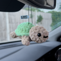 The Blossom Automotive Squishy Turtle - Car Mirror Hanger features a cute, plush turtle with a green shell and beige body, smiling as it hangs from your rearview mirror. Concrete wall and greenery appear outside the car window.